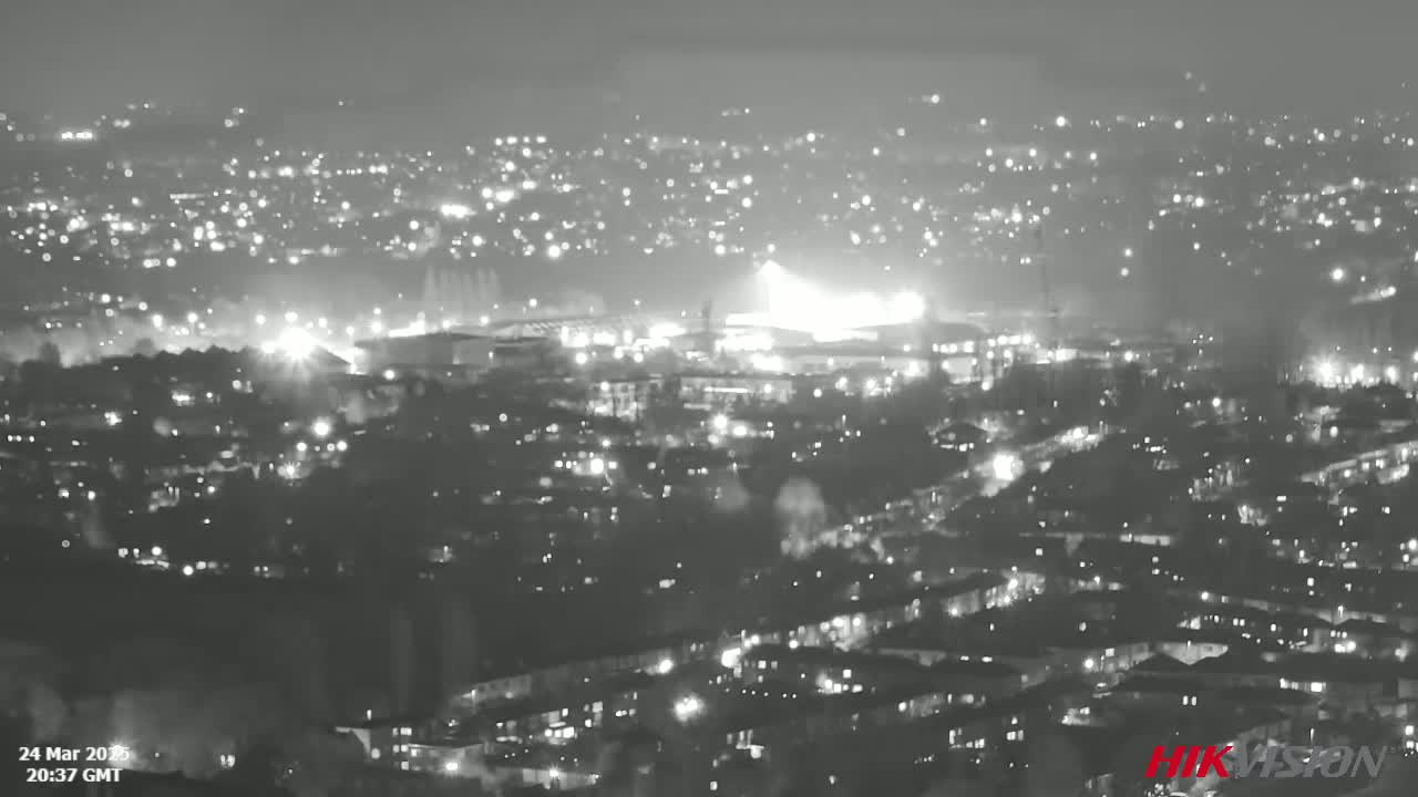 A nighttime, grayscale view of a city illuminated by numerous lights, with a brighter area possibly indicating a large event or industrial site in the mid-ground.