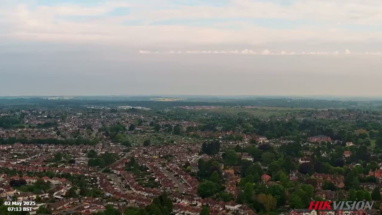 An aerial view shows a sprawling suburban area with many houses and green spaces under a mostly cloudy sky.
