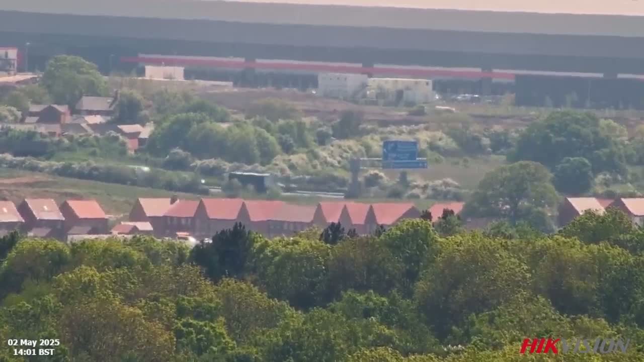 A long-range view shows a row of new red-roofed houses nestled among green trees, situated below a large industrial building and highway overpass on a hazy day.