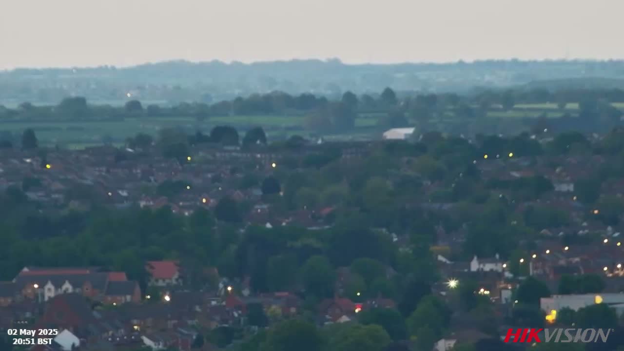 A hazy, twilight view shows a sprawling residential area with scattered lights, nestled amongst greenery and overlooking a distant, flat landscape.