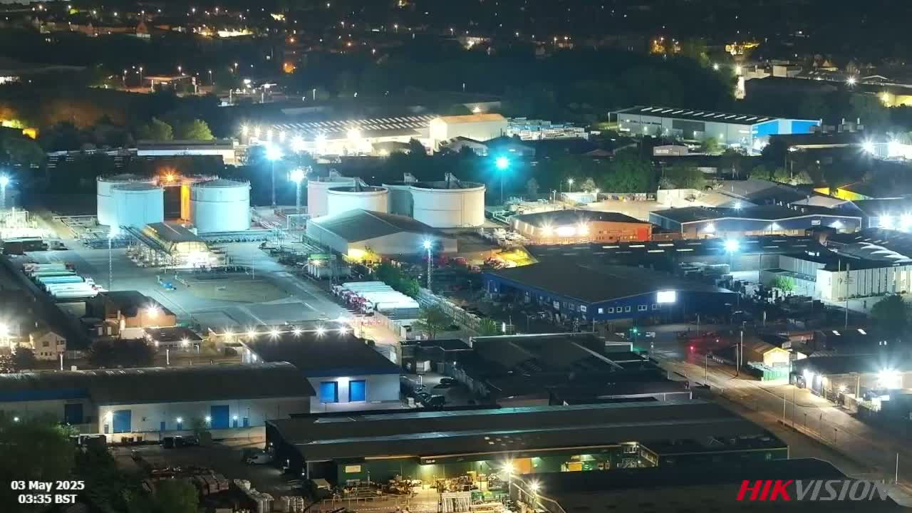 An industrial area at night, brightly lit, features large storage tanks and numerous buildings, under a clear sky.