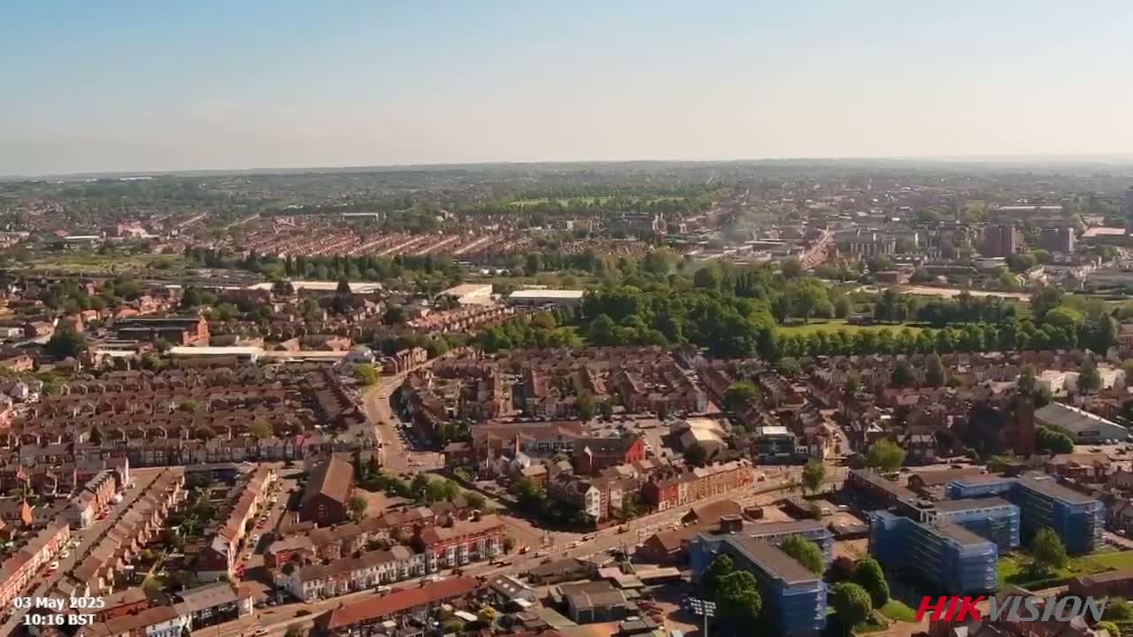 An aerial view shows a sprawling residential area with red-roofed houses, interspersed with green spaces and larger buildings under a clear, sunny sky.