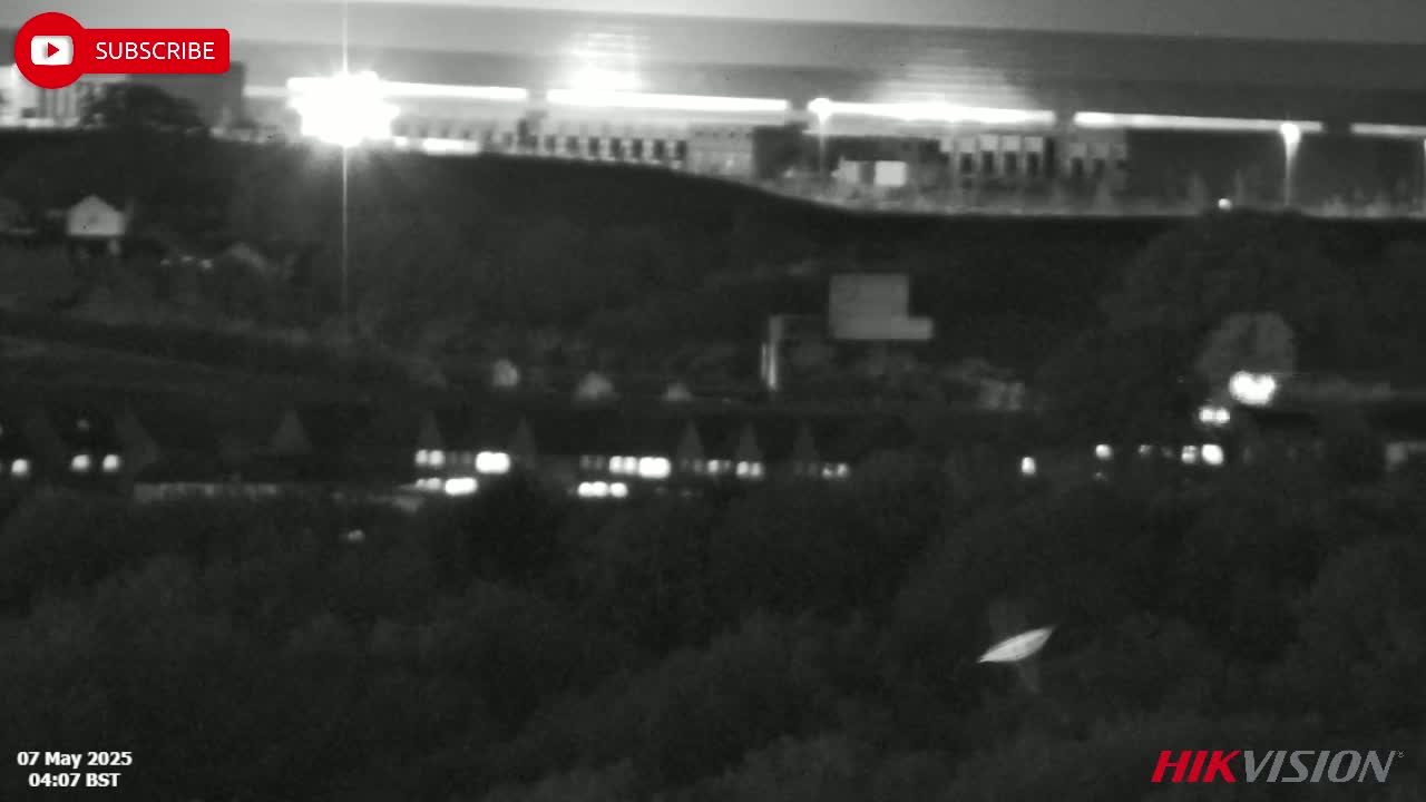 A nighttime, low-light image shows a dark landscape with sparsely lit buildings and vegetation, under a mostly clear sky.
