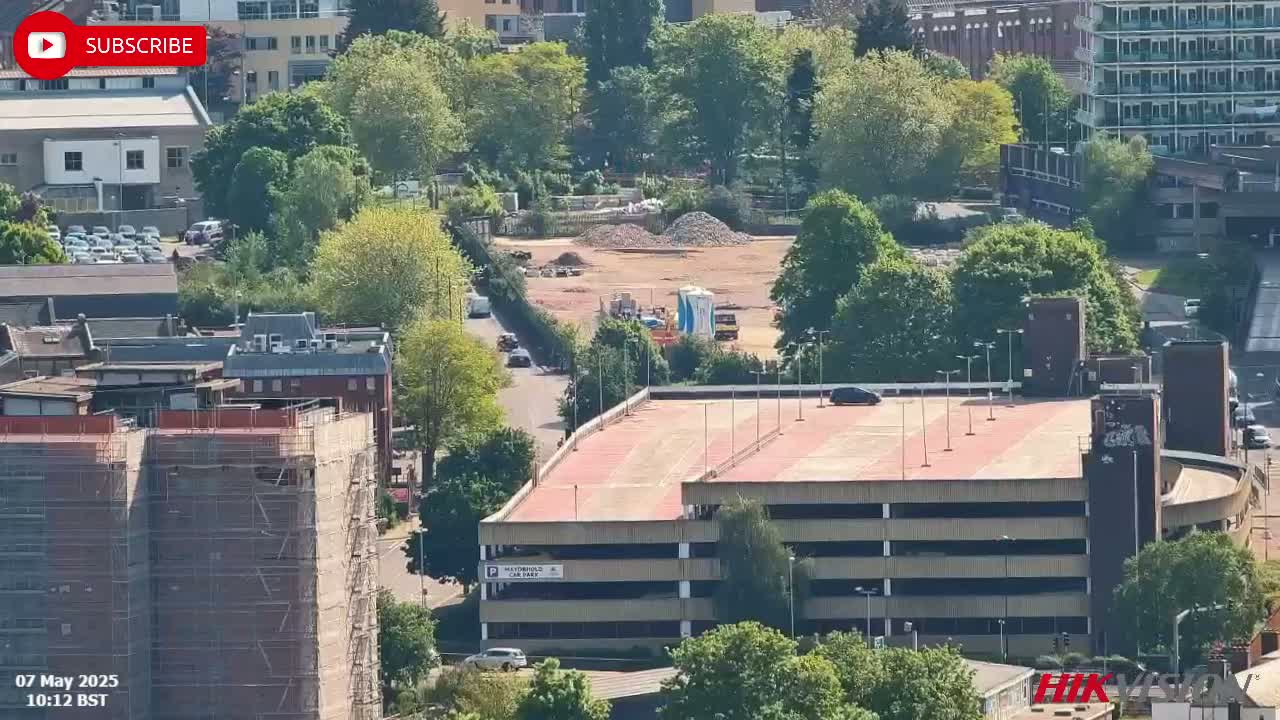 A high-angle, sunny view of a city landscape showing a multi-level parking garage, construction site, and surrounding buildings with lush green trees.