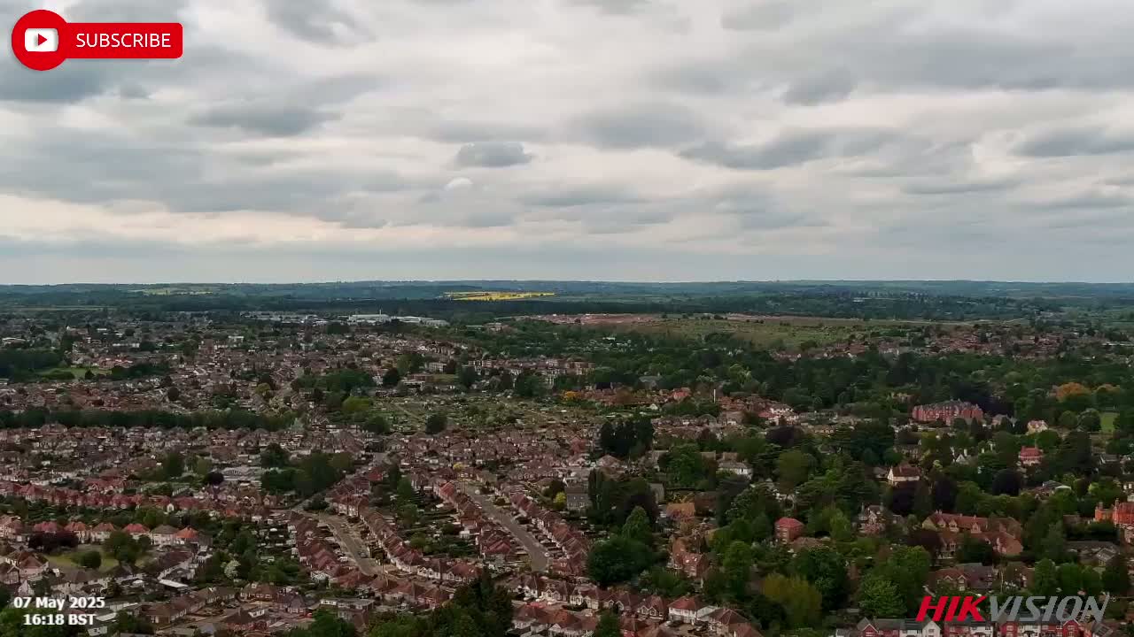 An aerial view shows a sprawling residential area with many houses and green spaces under a cloudy sky.