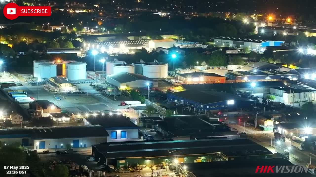 An aerial nighttime view shows brightly lit industrial buildings and storage tanks, surrounded by darker areas of trees and undeveloped land.
