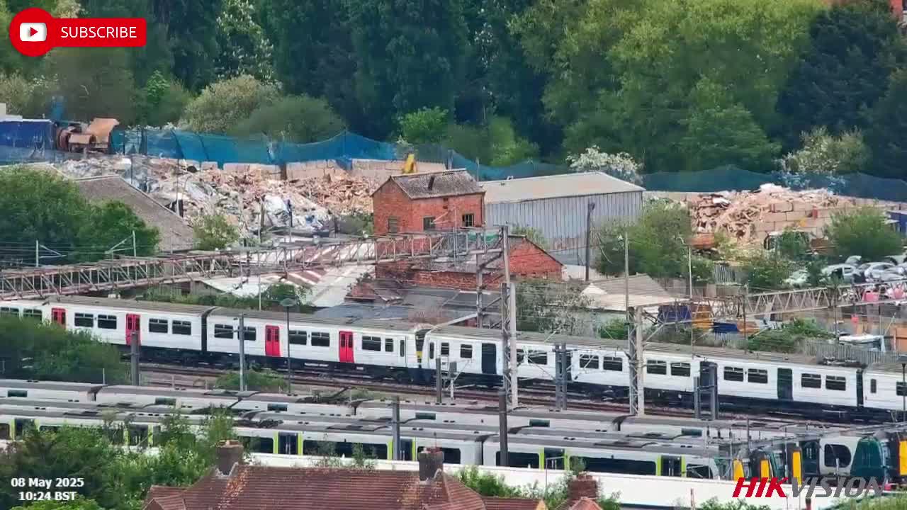 A commuter train travels past a demolition site on a sunny day, with several other trains parked nearby.