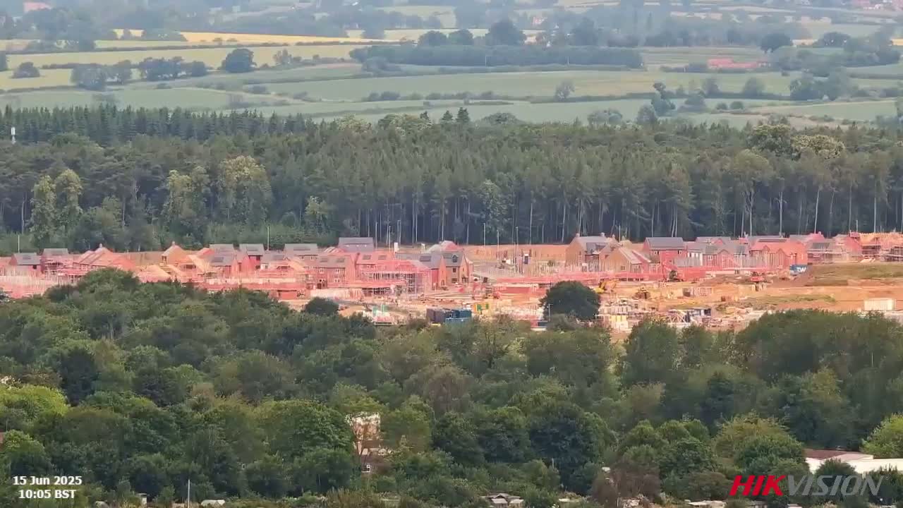 A large housing development under construction is nestled amongst a green forest, overlooking a vast, gently rolling landscape under a hazy sky.
