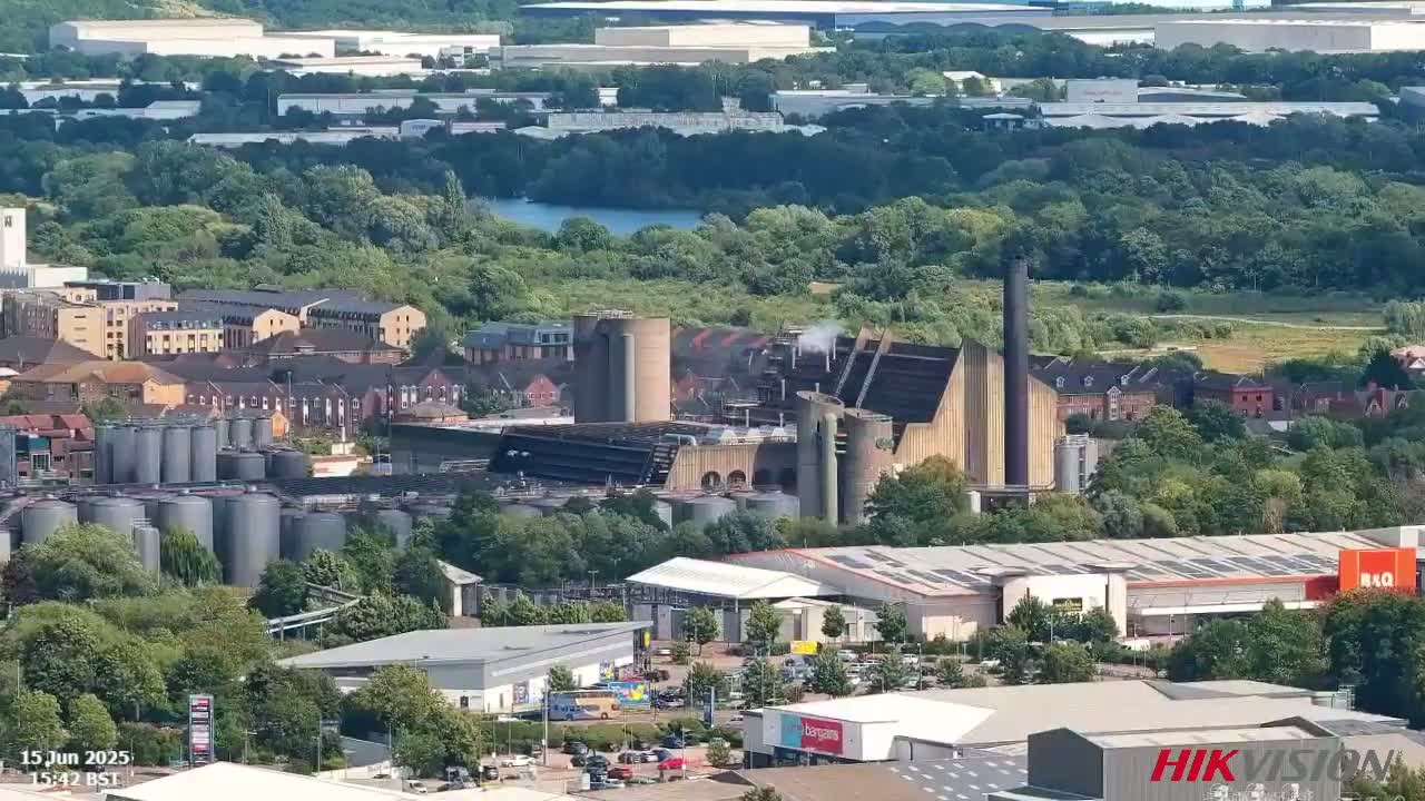 An aerial view shows an industrial area with large tanks, factories, and buildings surrounded by greenery on a sunny day.