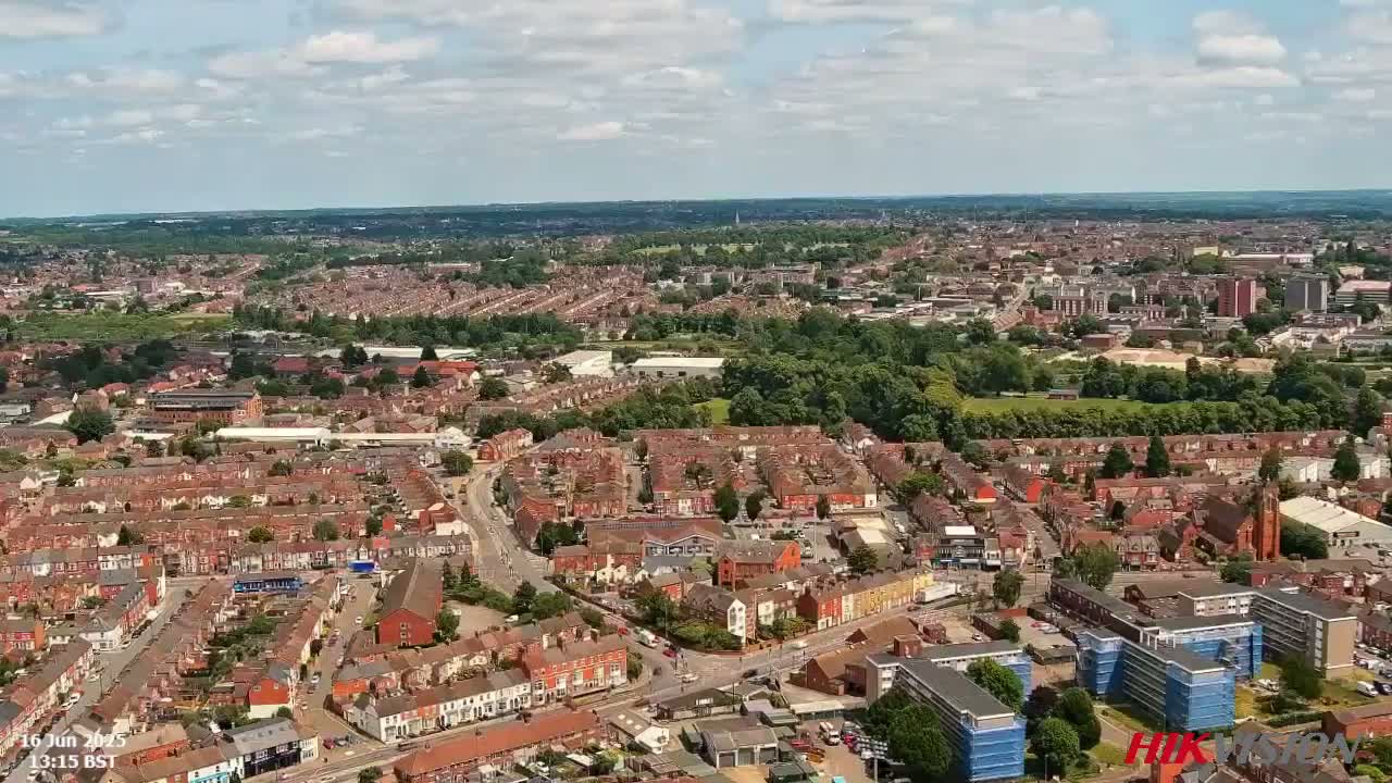 An aerial view shows a sprawling residential area with red-brick houses, interspersed with green spaces and taller buildings, under a partly cloudy sky.