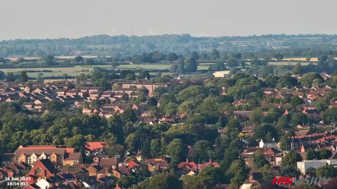 A sprawling residential area with many houses and trees, situated in a landscape featuring fields and distant wind turbines under a hazy sky.