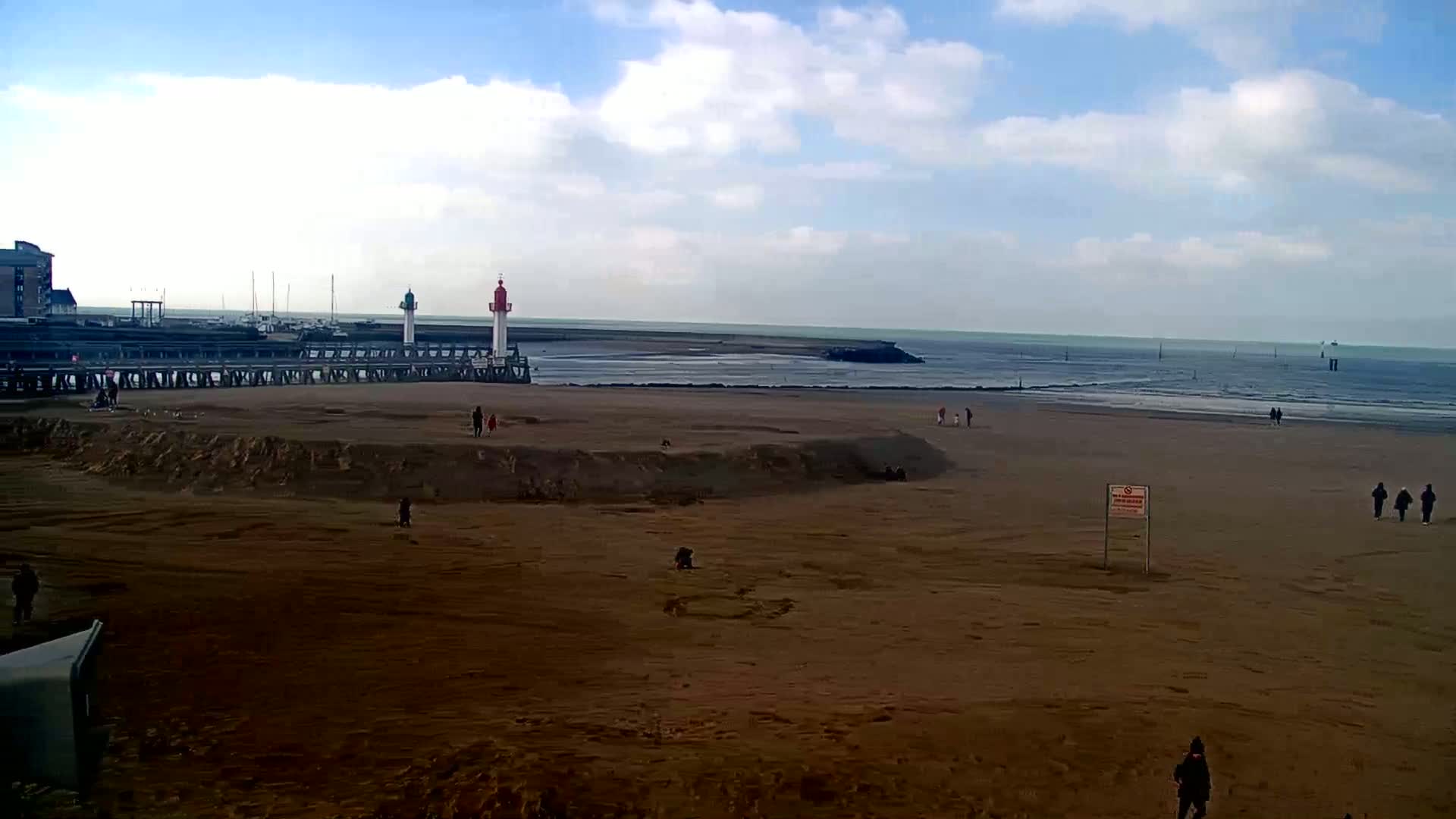 A mostly sunny day shows a sandy beach with a few people walking, a pier with two lighthouses, and calm ocean waters at low tide.