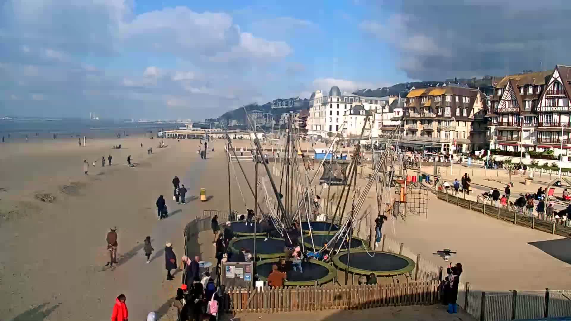 A sandy beach with people walking and playing on trampolines, overlooking a seaside town under partly cloudy skies.