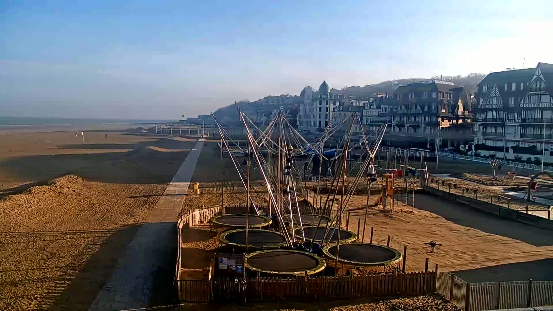 A sandy beach with a boardwalk and a cluster of trampolines near buildings on a clear, sunny day.