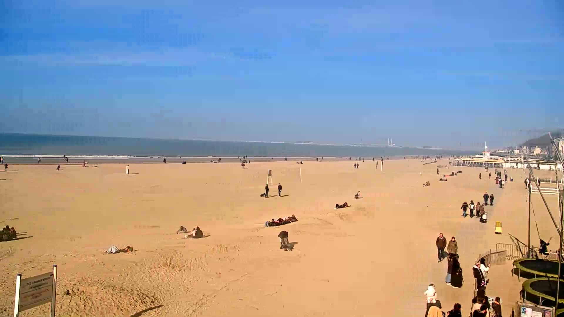 A wide sandy beach with scattered people sunbathing and walking, under a clear blue sky, and a calm ocean in the background.