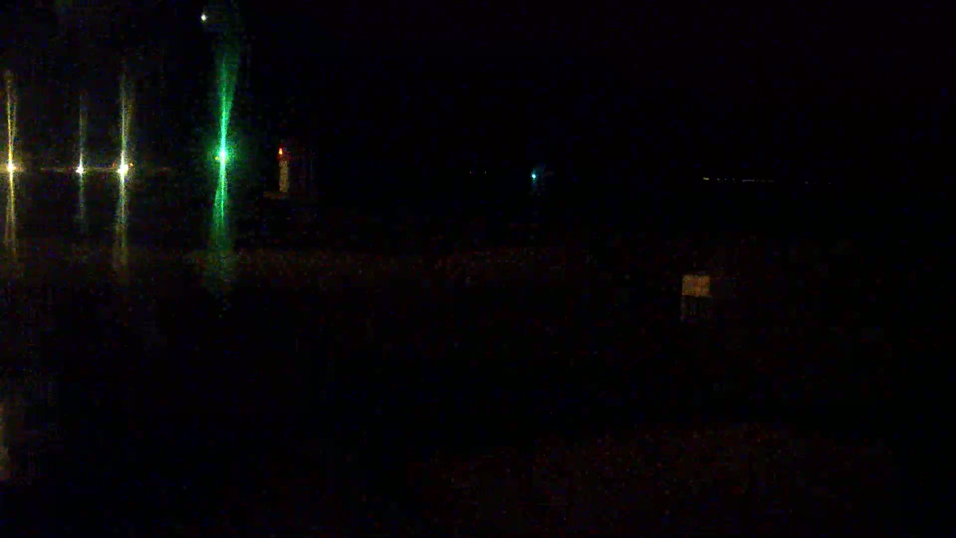A dark, nighttime scene shows a few faint lights in the distance and reflections of lights on a wet surface in the foreground.