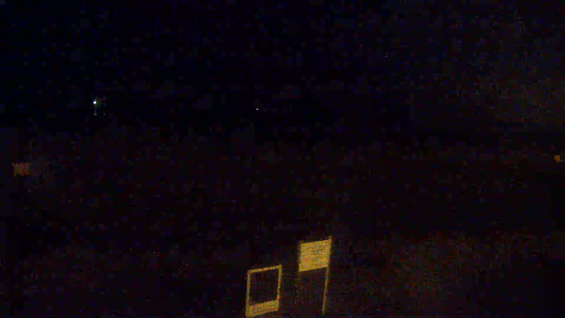 A nighttime outdoor scene shows a dark, blurry landscape with a few faint lights in the distance and a blurred, illuminated square shape in the bottom right corner.