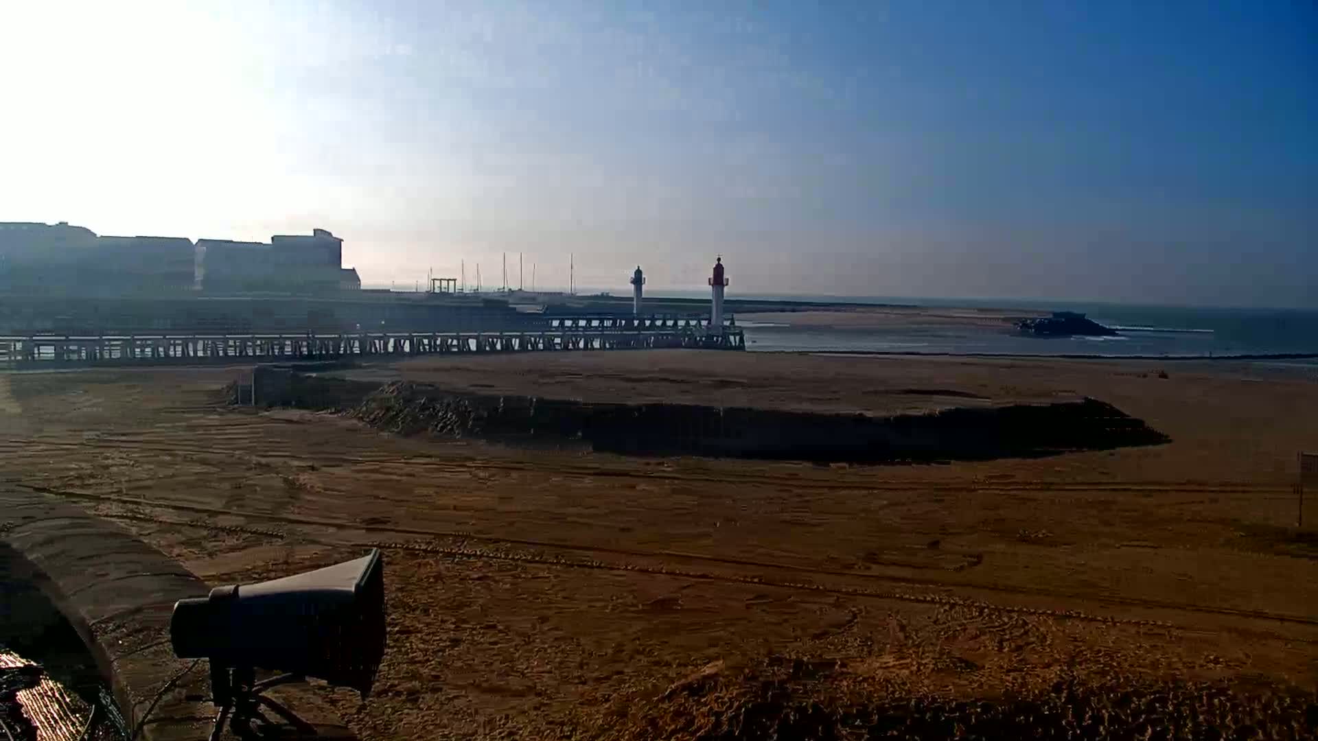 A sandy beach stretches out before a pier with two lighthouses, a calm sea visible in the background under a clear, bright sky.