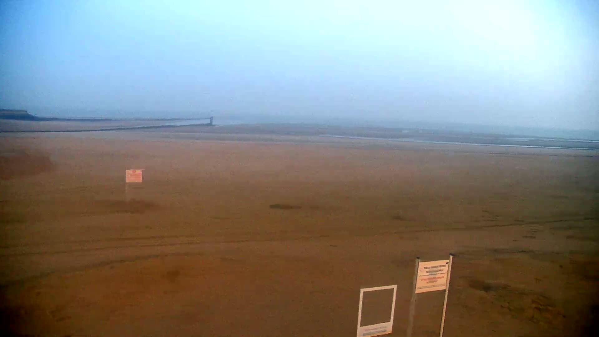 A hazy, expansive sandy beach stretches to a distant, indistinct shoreline under a pale sky.