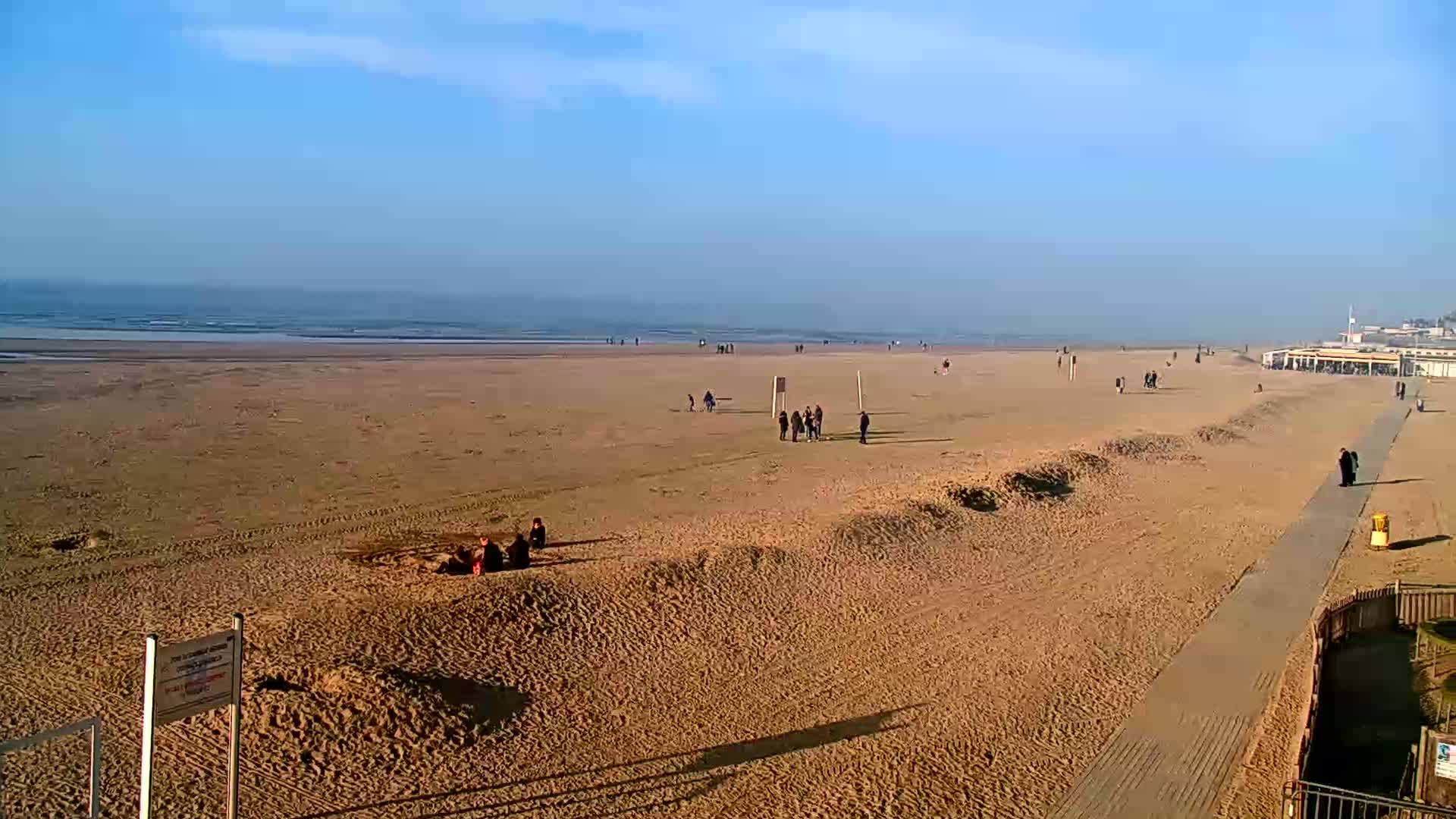A wide sandy beach with a few people scattered across it stretches to a calm, light-blue ocean under a clear, bright sky.