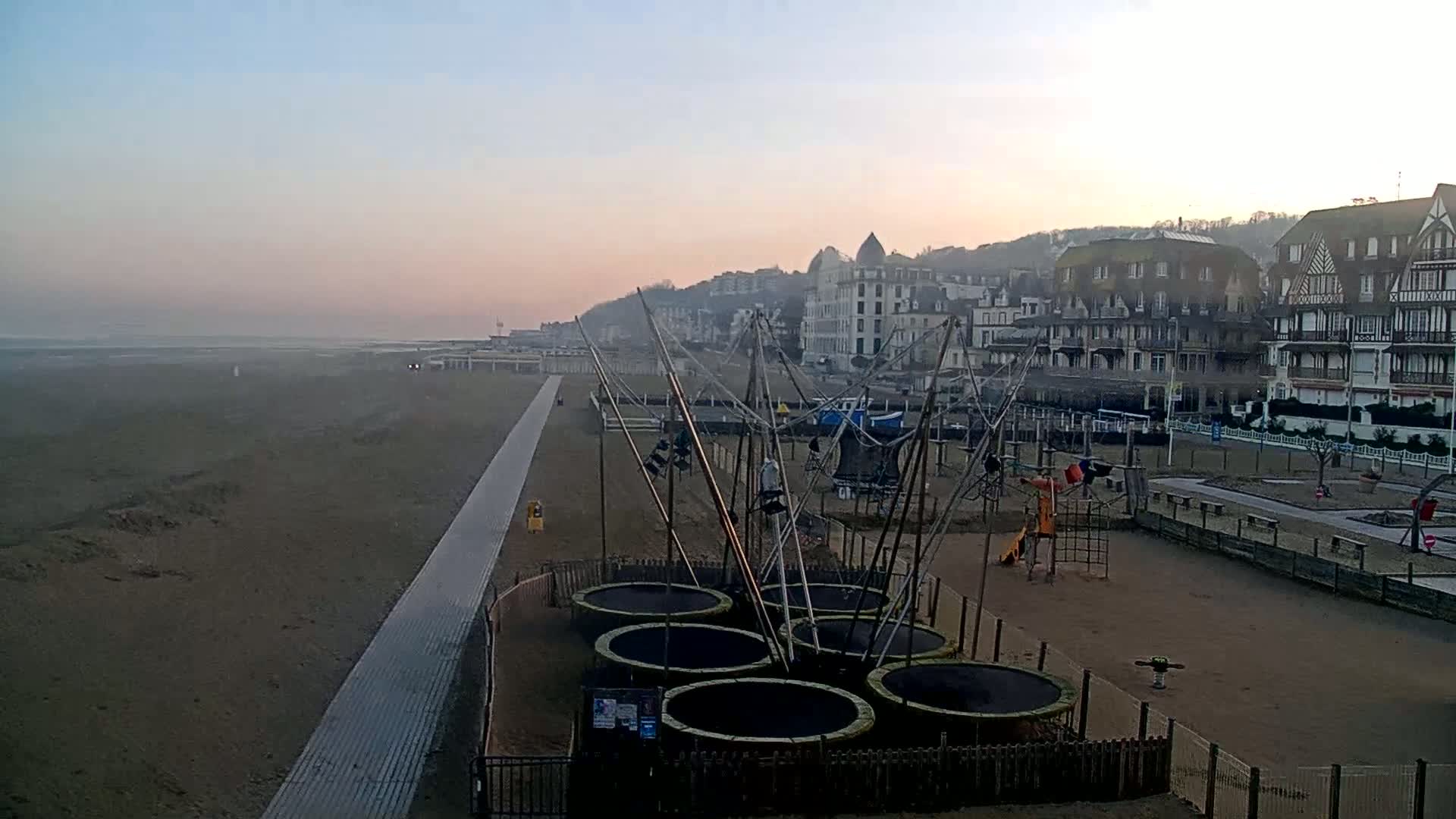 A hazy sunrise illuminates a beach with a boardwalk, playground, and several trampolines near a row of buildings on a hillside.