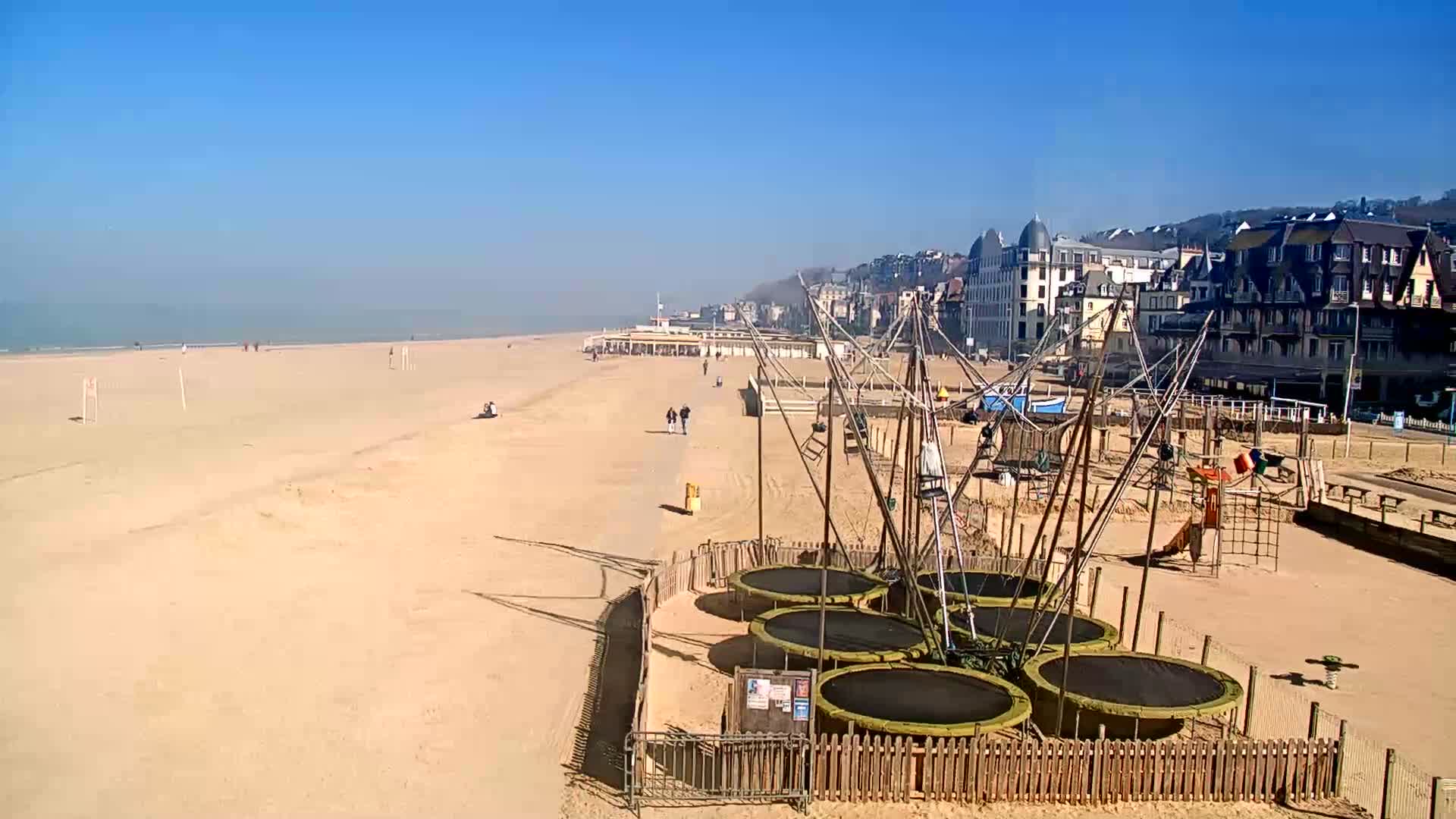 A sandy beach with several trampolines and a playground is adjacent to a row of buildings under a clear blue sky.