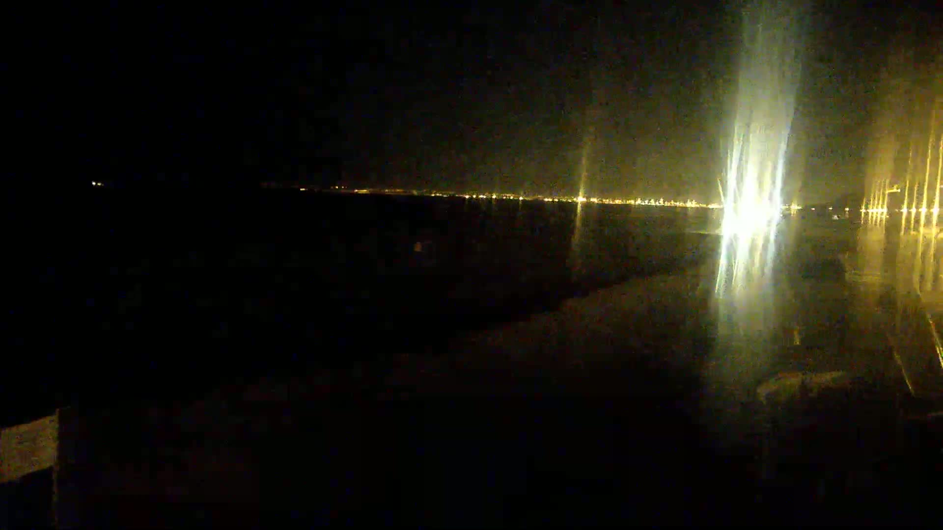 A dark, nighttime scene shows a distant city skyline illuminated by lights reflected on a dark, wet surface.