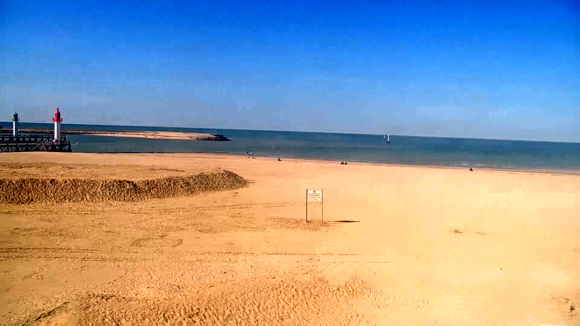 A sandy beach stretches out to a calm sea under a clear blue sky, with a small pier and lighthouses visible in the distance.