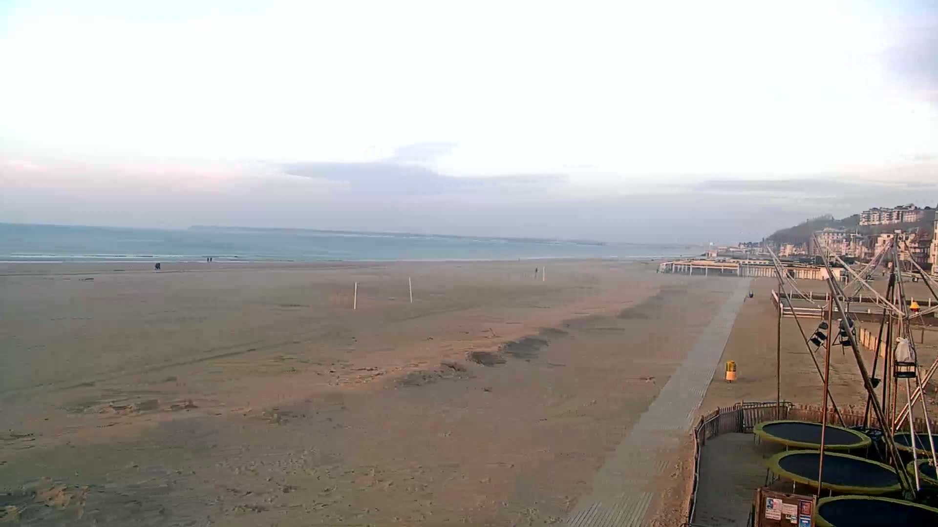 A wide sandy beach stretches to a calm ocean under an overcast sky, with a boardwalk and playground equipment visible in the foreground.