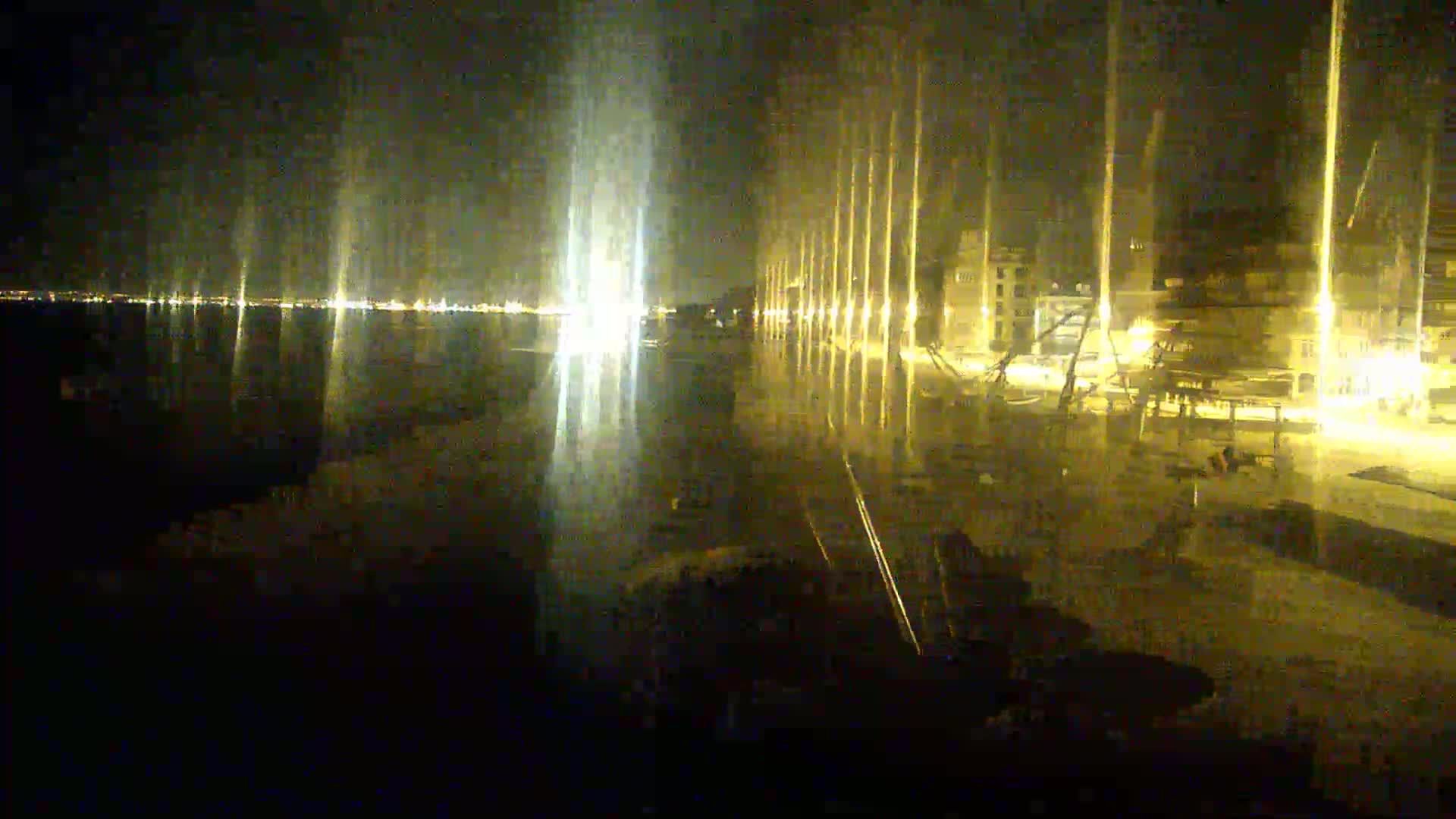A nighttime waterfront scene shows a city's lights reflected on calm water, with tall, illuminated poles lining a paved area.