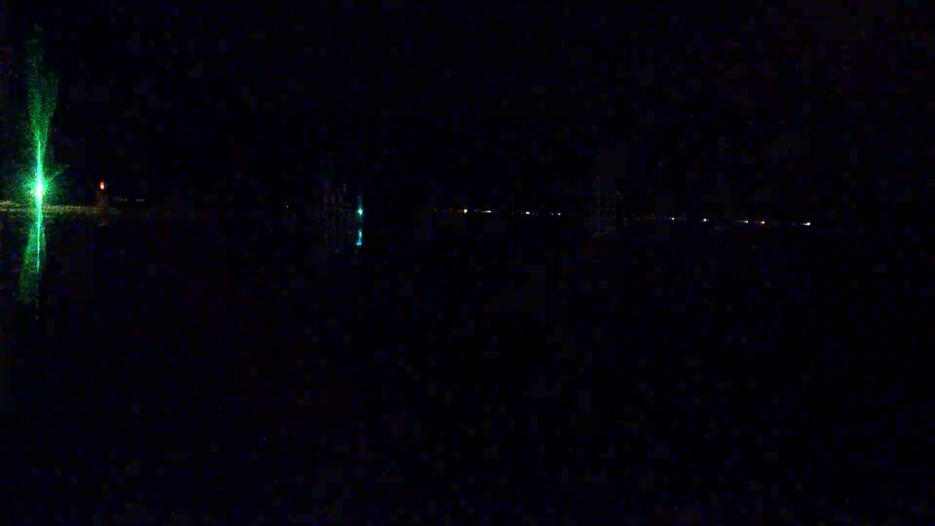 A dark, nighttime scene shows a body of water reflecting a few distant lights and a bright green light, possibly from a navigational aid, closer to the viewer.