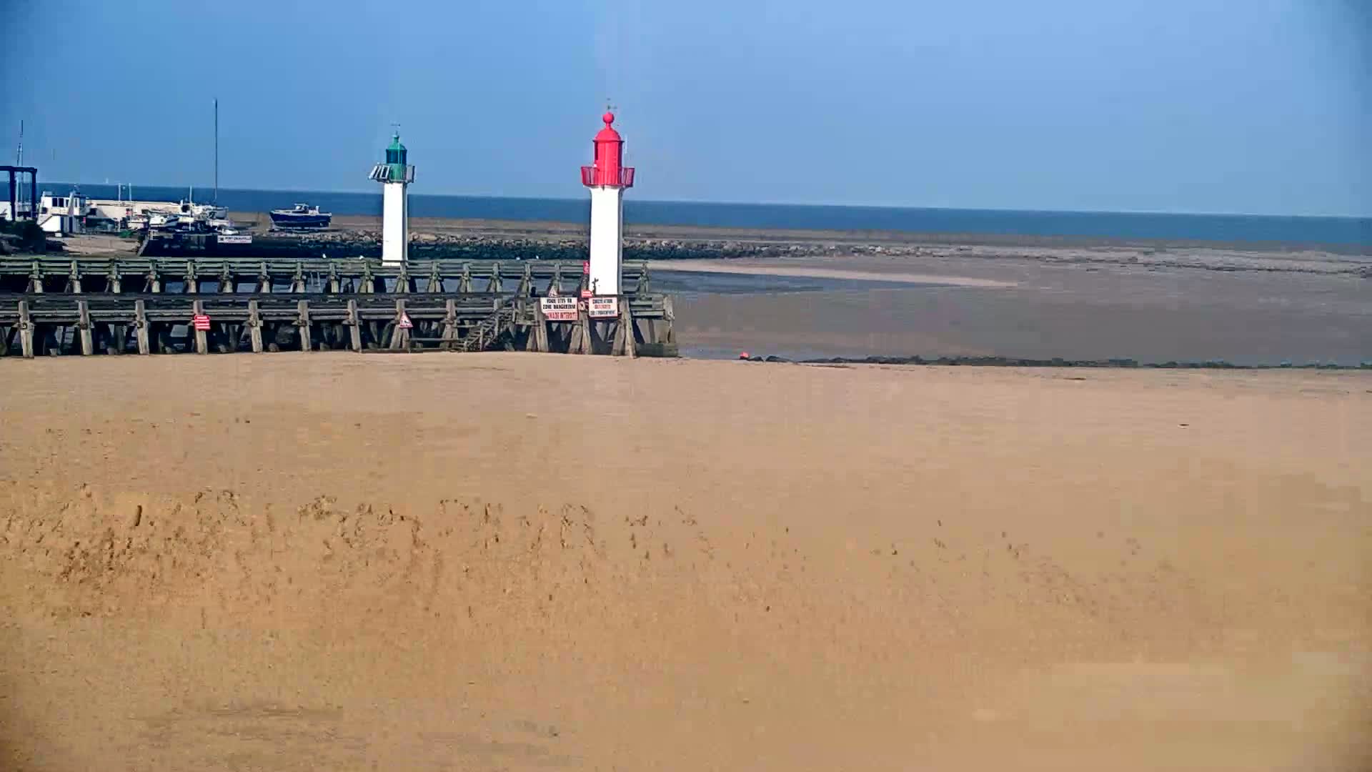 A sandy beach stretches out to a calm ocean under a clear blue sky, with two small lighthouses standing on a wooden pier behind it.