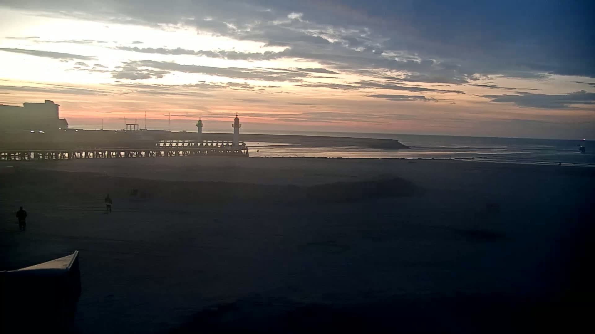 A beach at sunset with two lighthouses visible on a pier, under a partly cloudy sky.