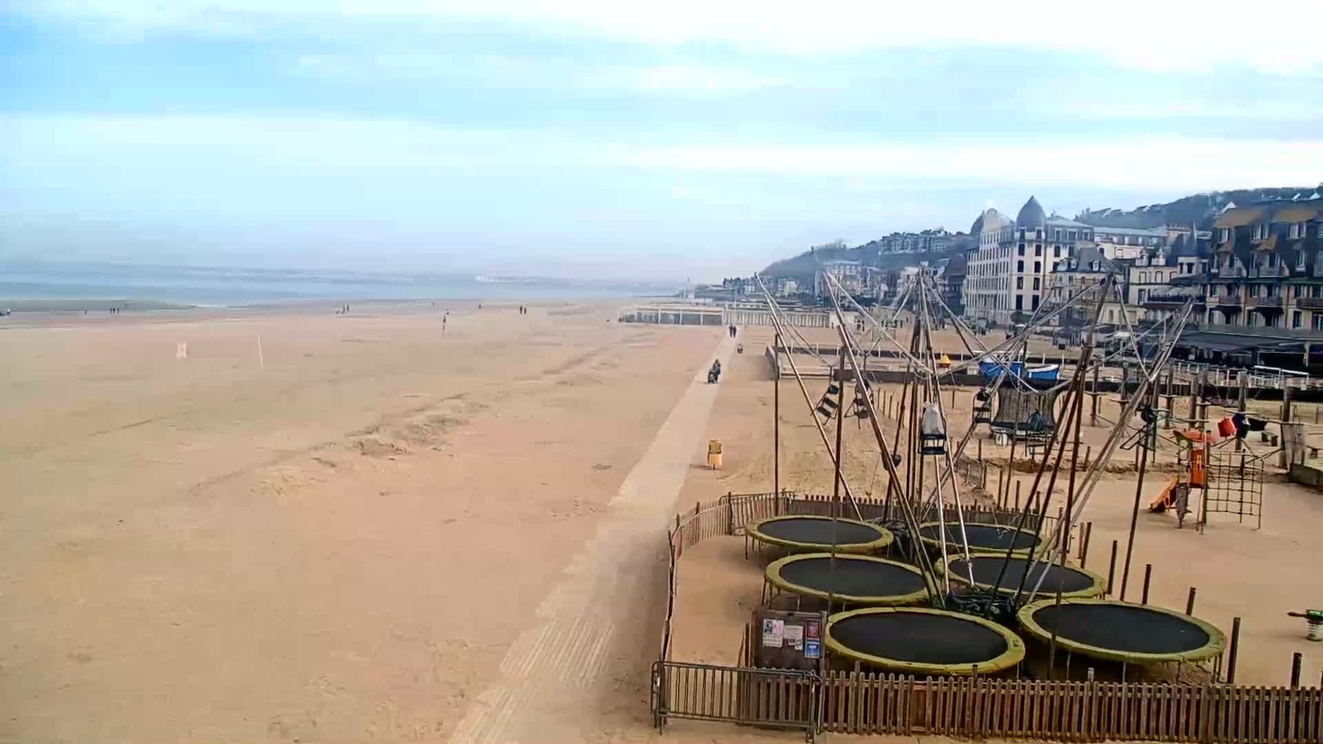 A sandy beach with a boardwalk, several trampolines, and a playground is visible, with a calm ocean and buildings in the background under a mostly cloudy sky.