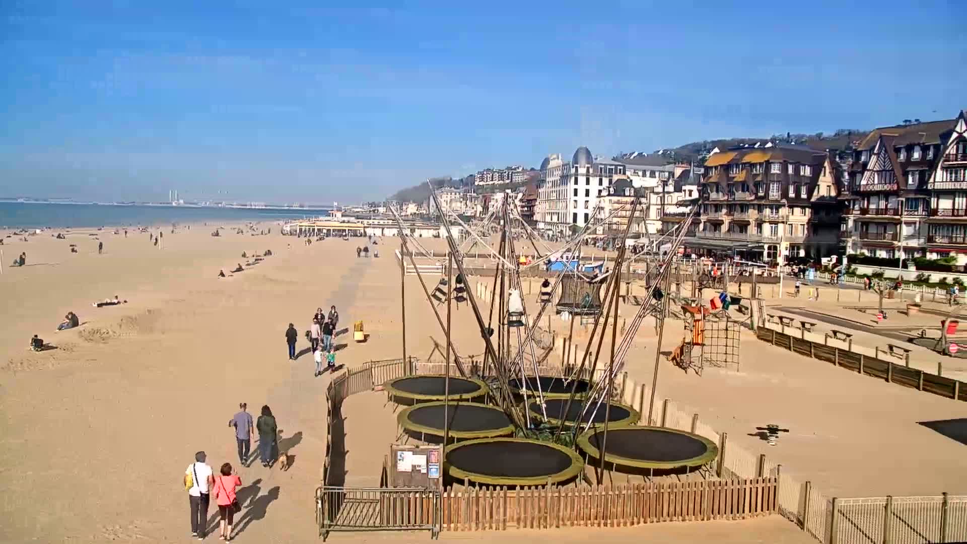 A sandy beach with people sunbathing and walking, featuring a playground with trampolines, is bordered by buildings under a clear blue sky.