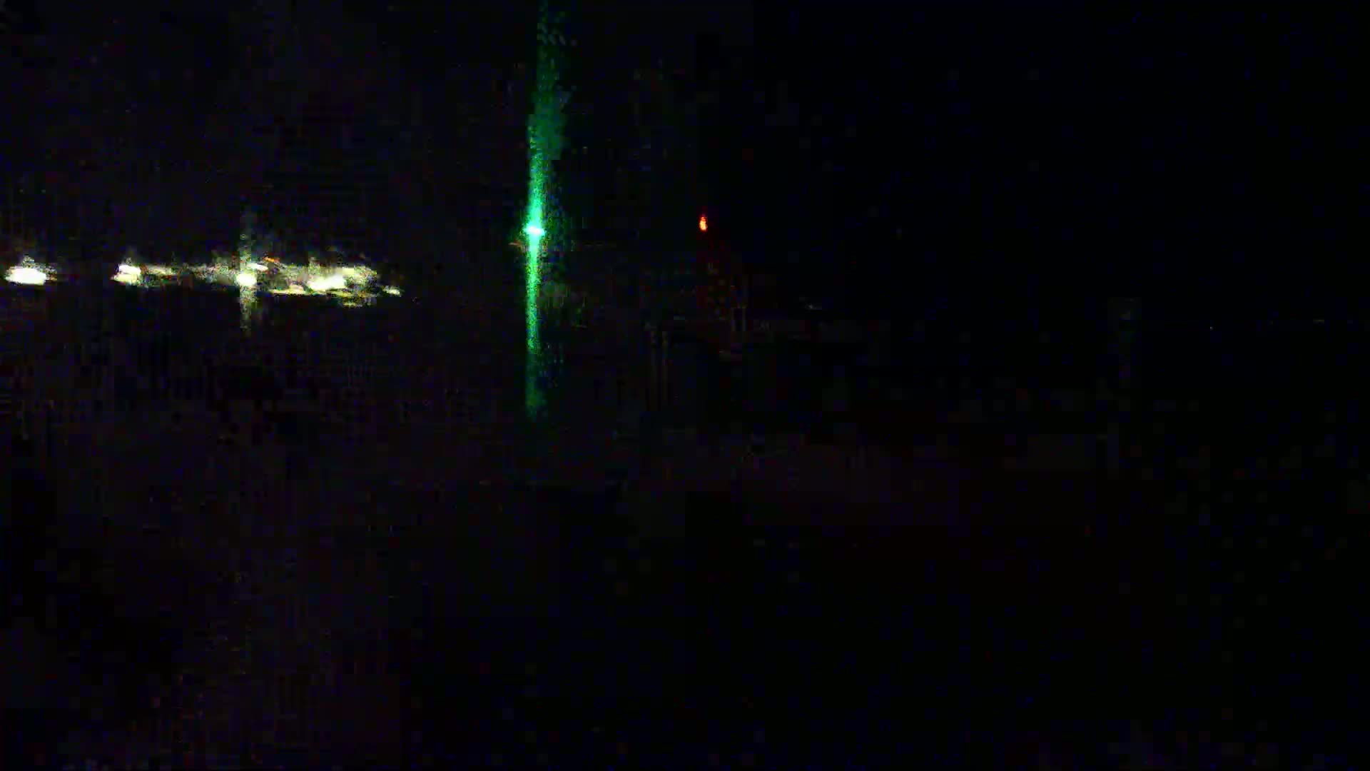 At night, a tall, bright green light is visible next to a smaller, faint orange light, with several other small lights in the distance.