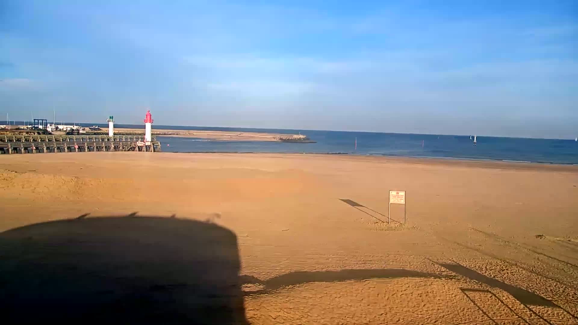A sandy beach stretches out to a calm ocean under a clear blue sky, with a wooden pier and two lighthouses visible in the distance.