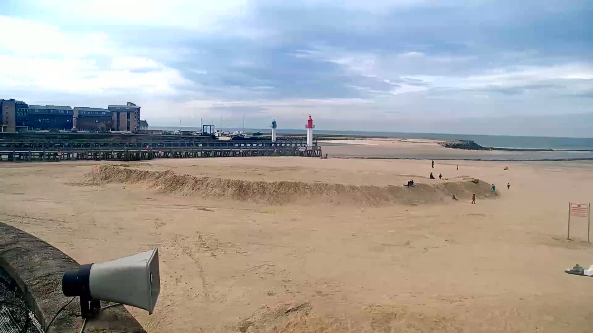 A mostly empty sandy beach stretches out to a calm sea under a cloudy sky, with a pier, buildings, and two lighthouses visible in the background.