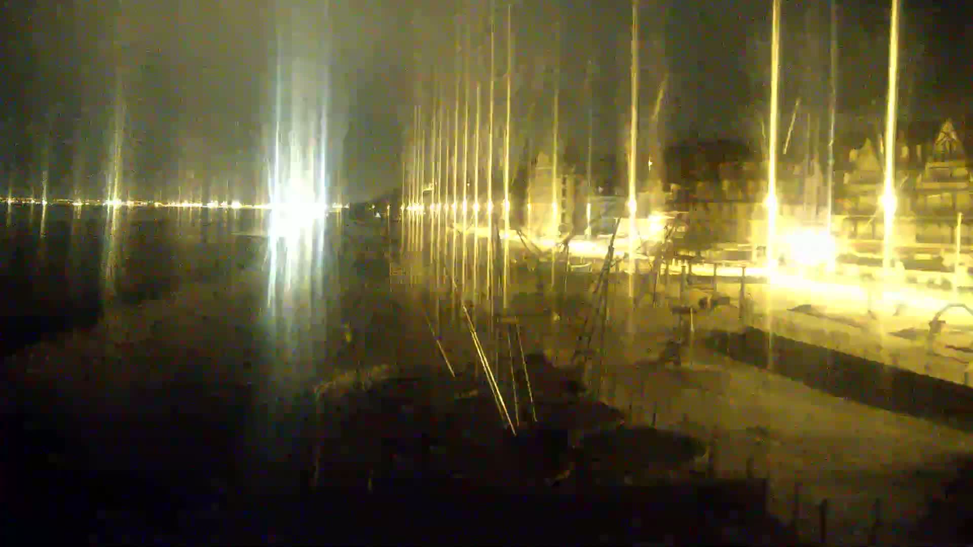 At night, a body of water reflects the lights of a town's waterfront, where numerous tall poles stand along a dock.