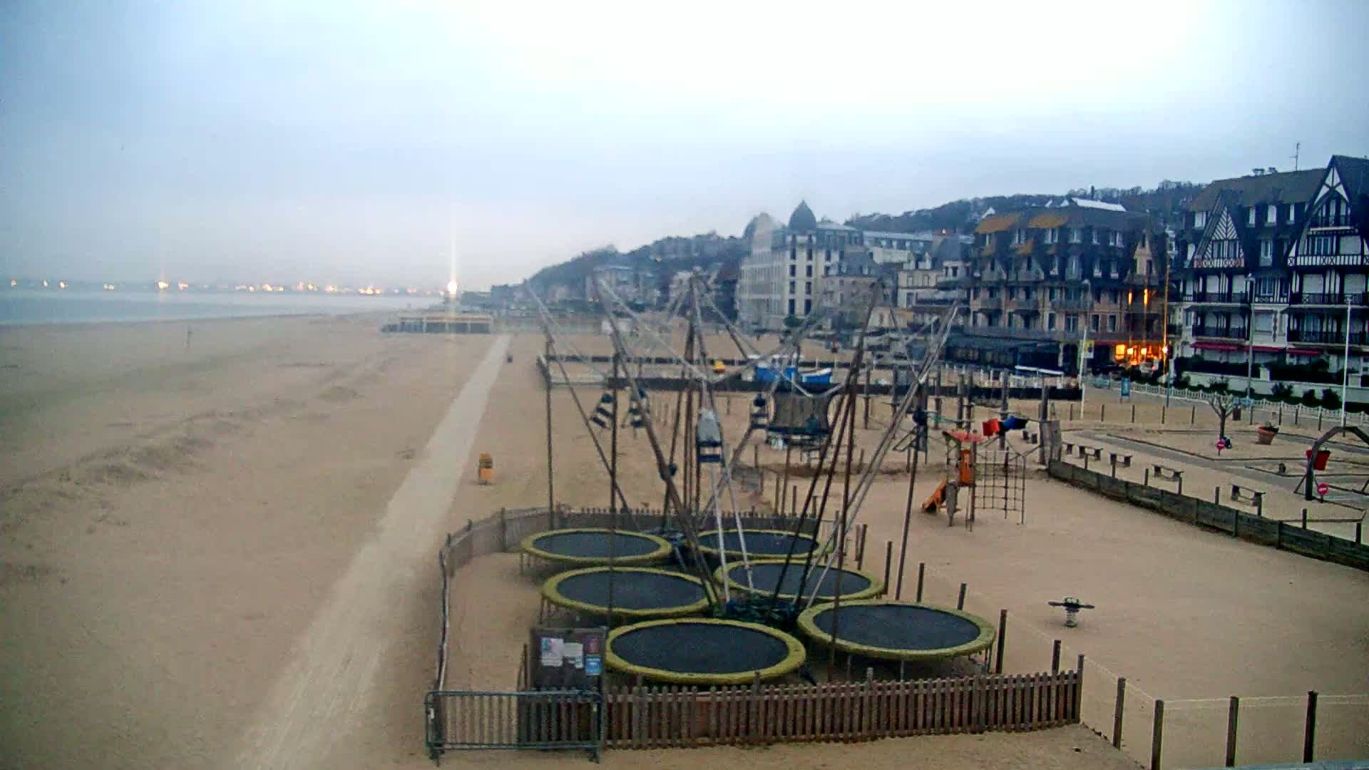 A sandy beach with a playground featuring several trampolines is situated near a row of buildings under an overcast sky.