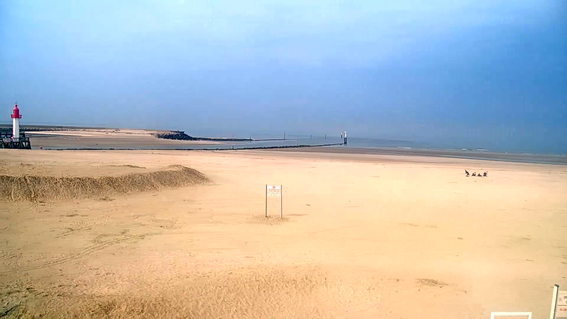 A mostly empty sandy beach stretches to a calm ocean under a clear blue sky, with a small lighthouse and a few people visible in the distance.