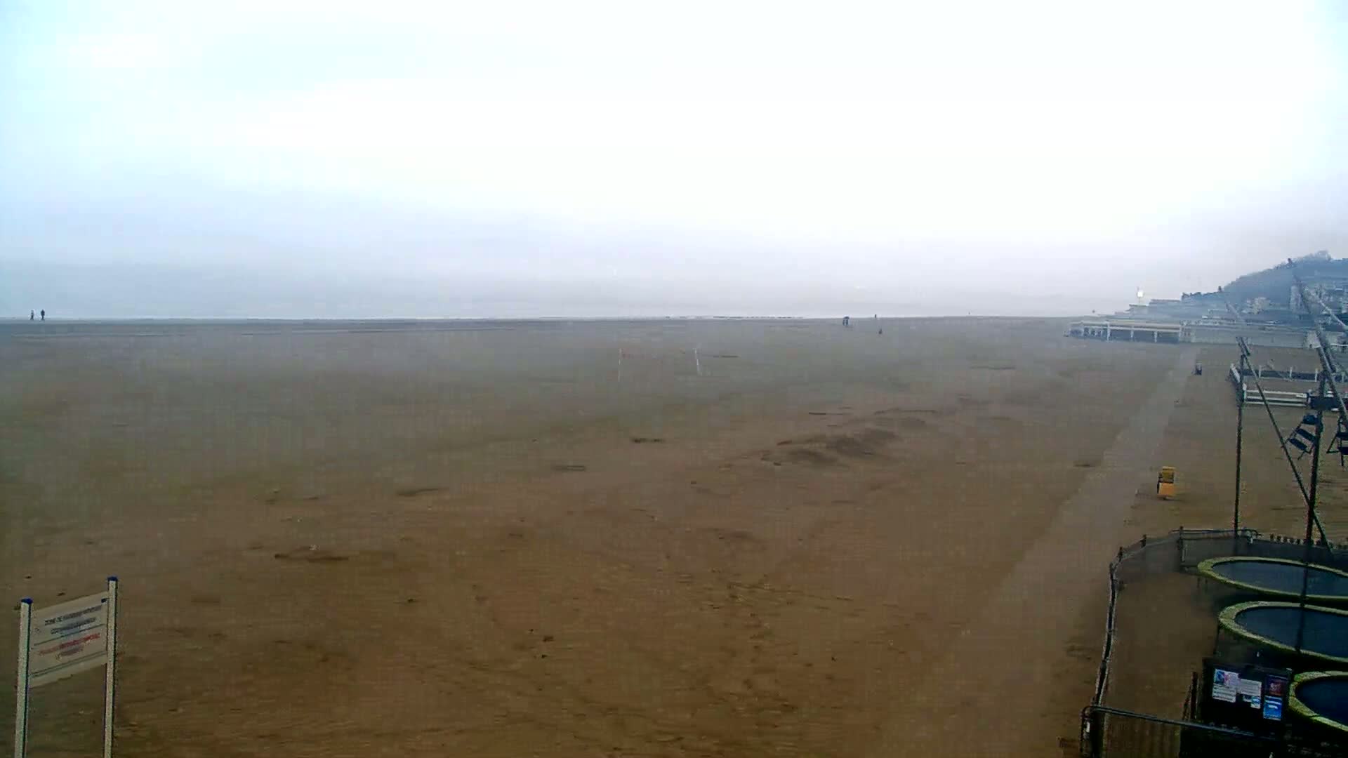 A wide, flat sandy beach stretches to a hazy ocean under a cloudy sky, with a few people visible in the distance and some beachside structures to the right.