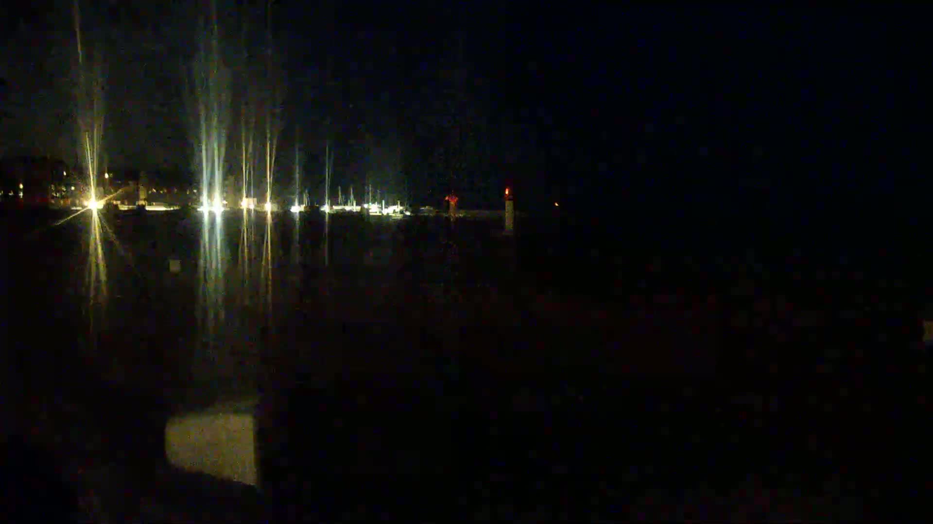 At night, the lights of boats in a harbor reflect on dark water, with a faint light visible in the distance.