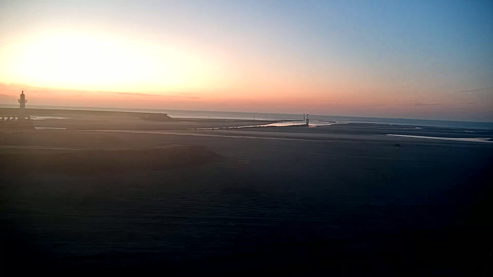 A coastal scene at sunset shows a lighthouse, a calm sea, and a vast expanse of dark sand.