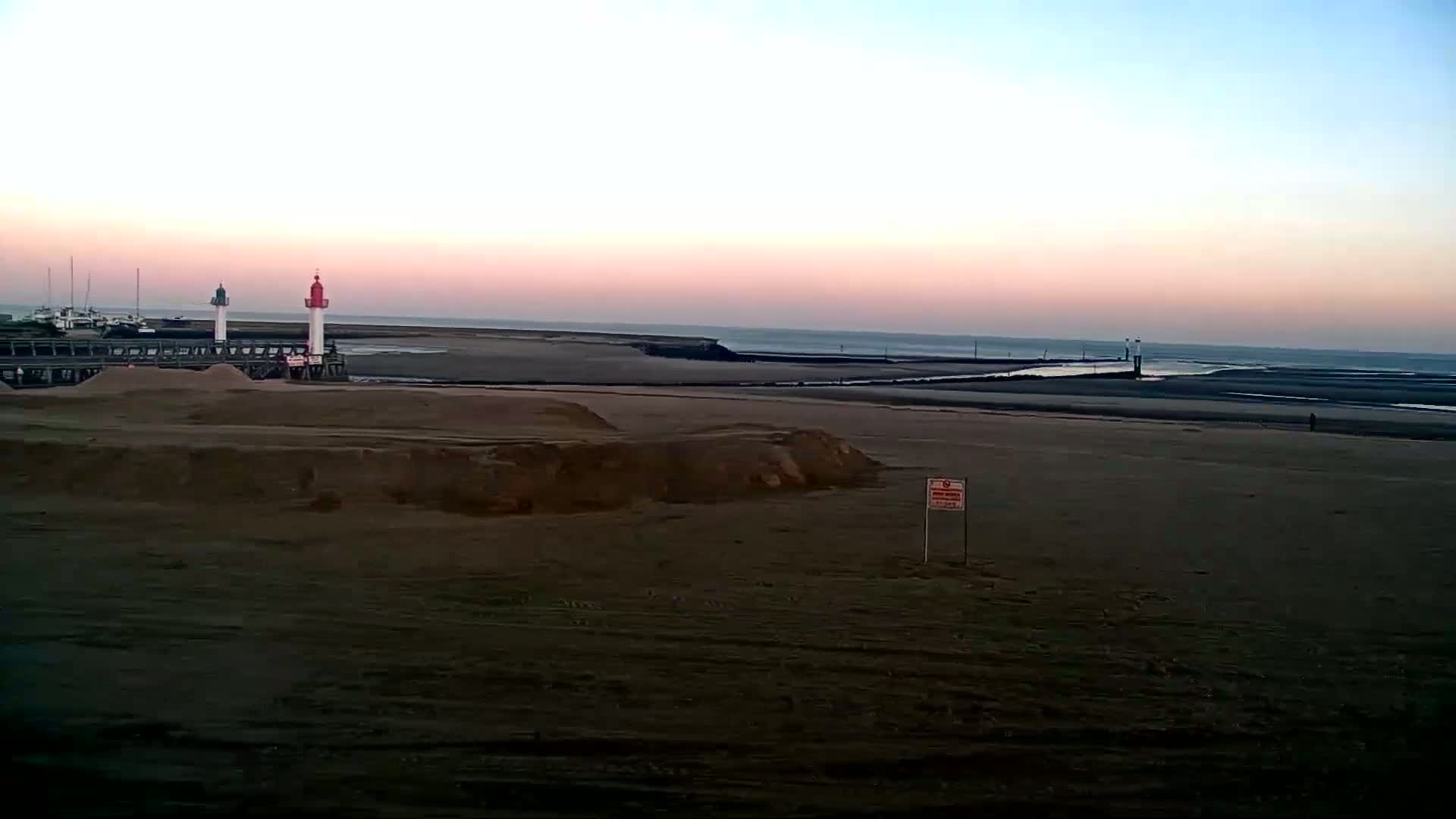 A sandy beach at sunset shows two small lighthouses on a pier in the distance, with calm water and a flat horizon.