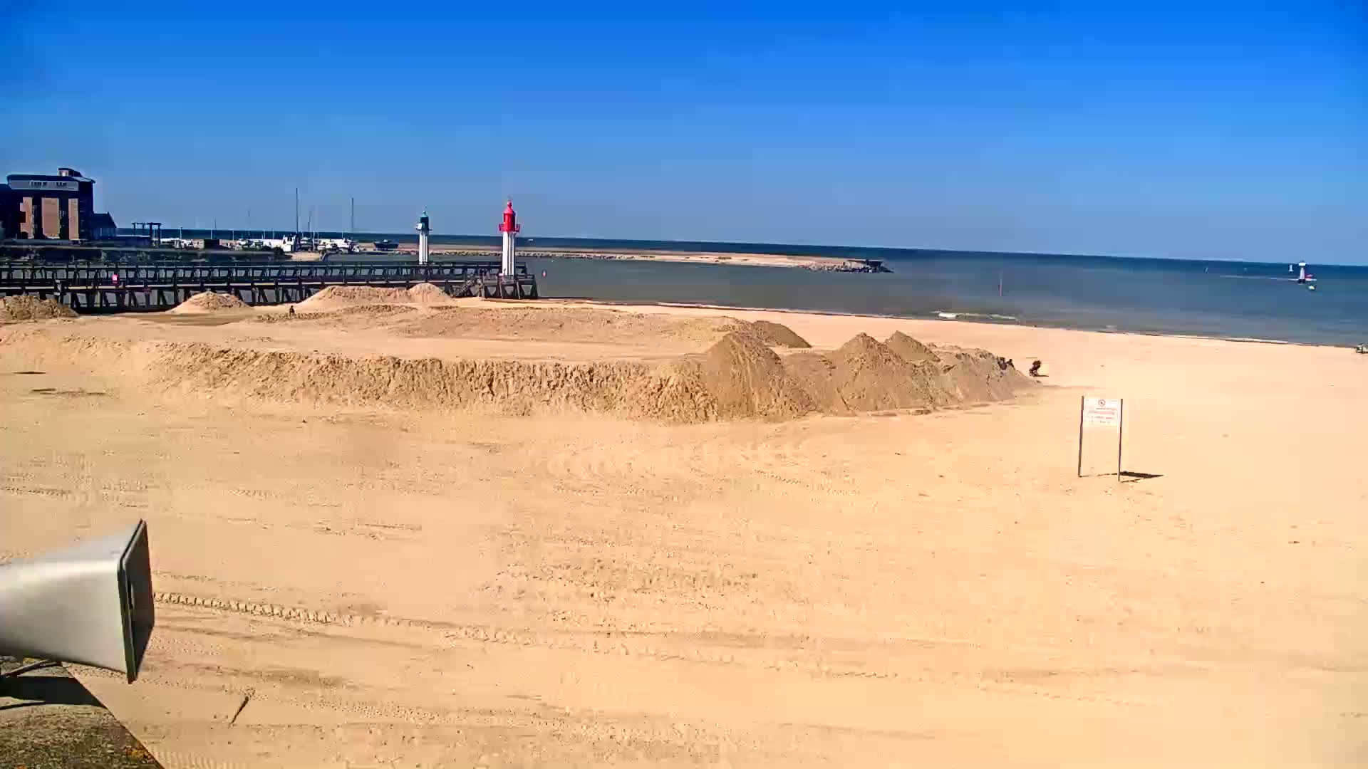 A sandy beach with piles of sand, a lighthouse, and calm blue water under a clear sky.