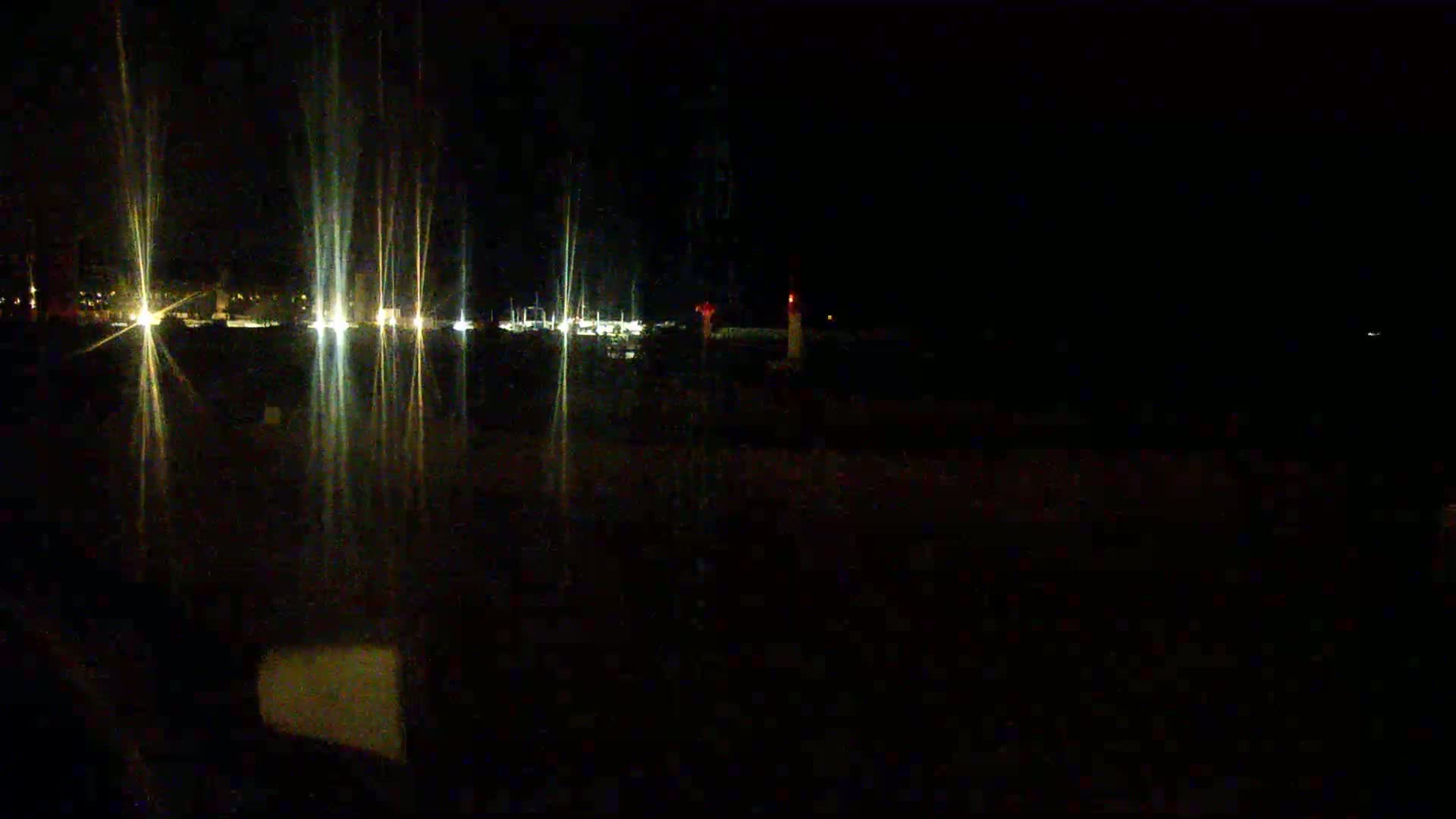 At night, bright lights from a distant town are visible across a dark body of water, with a single red light in the distance.