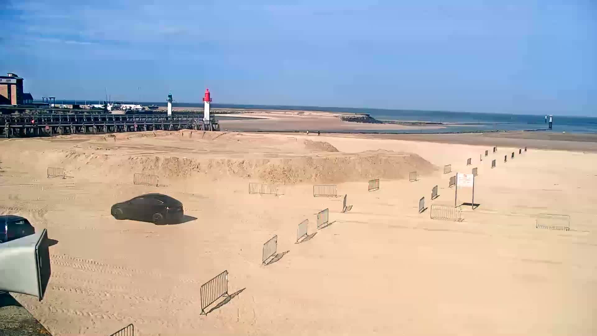 A sandy beach with a car and several short metal barriers stretches out to a calm sea under a clear blue sky.
