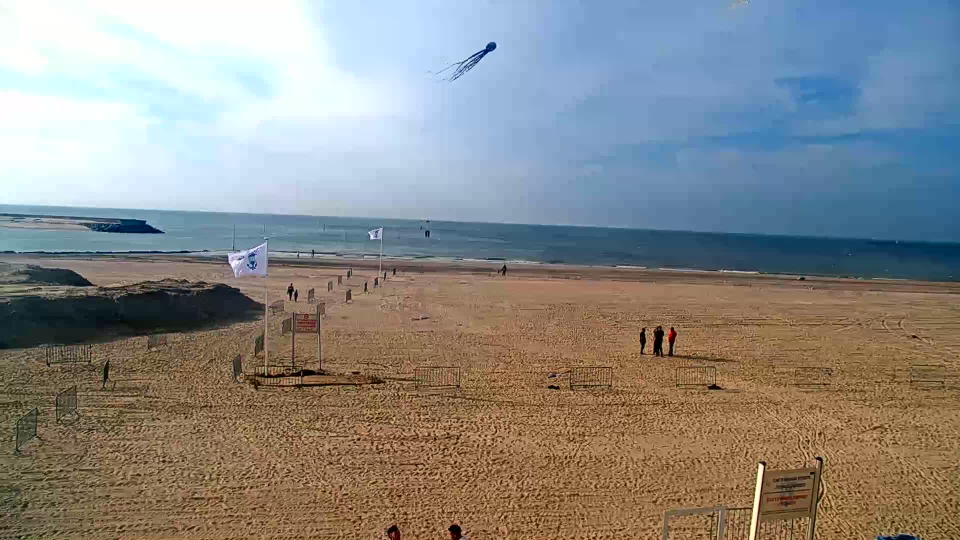 A sandy beach with a few people and a kite in the sky on a partly cloudy day, overlooking a calm sea.