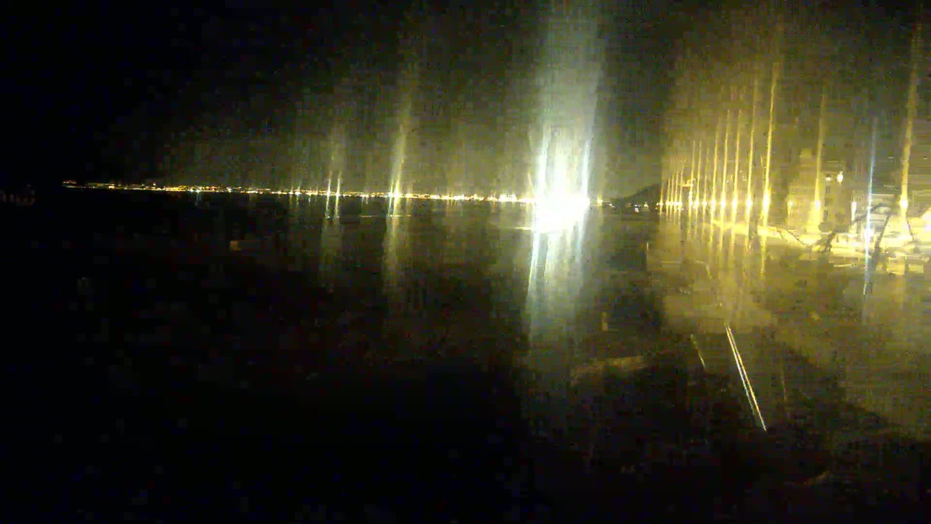 A nighttime view of a body of water reflecting the lights of a city across the way, with illuminated columns or structures lining the shore in the foreground.